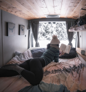 A person lying on a bed reading a book inside a Karma campervan.